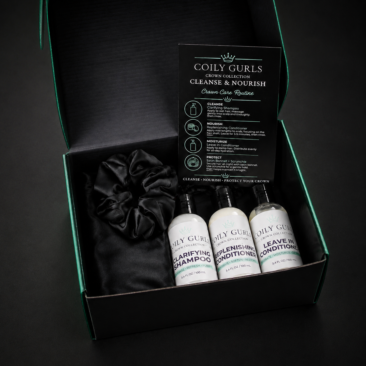 Coily Gurls Brown Collective hair care set in a black gift box, featuring three white bottles of shampoo, conditioner, and leave-in conditioner, a black satin scrunchie, and an instruction card, set against a dark background.